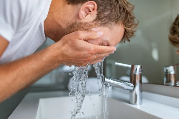 Kobieta w białym ręczniku na głowie myje twarz w łazience.